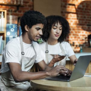 A black man and woman, both wearing the same apron, work at a table while looking at a computer and reciepts.